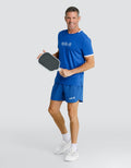 A smiling man wearing a blue athletic t-shirt and matching shorts, holding a paddle, standing against a light gray studio background.